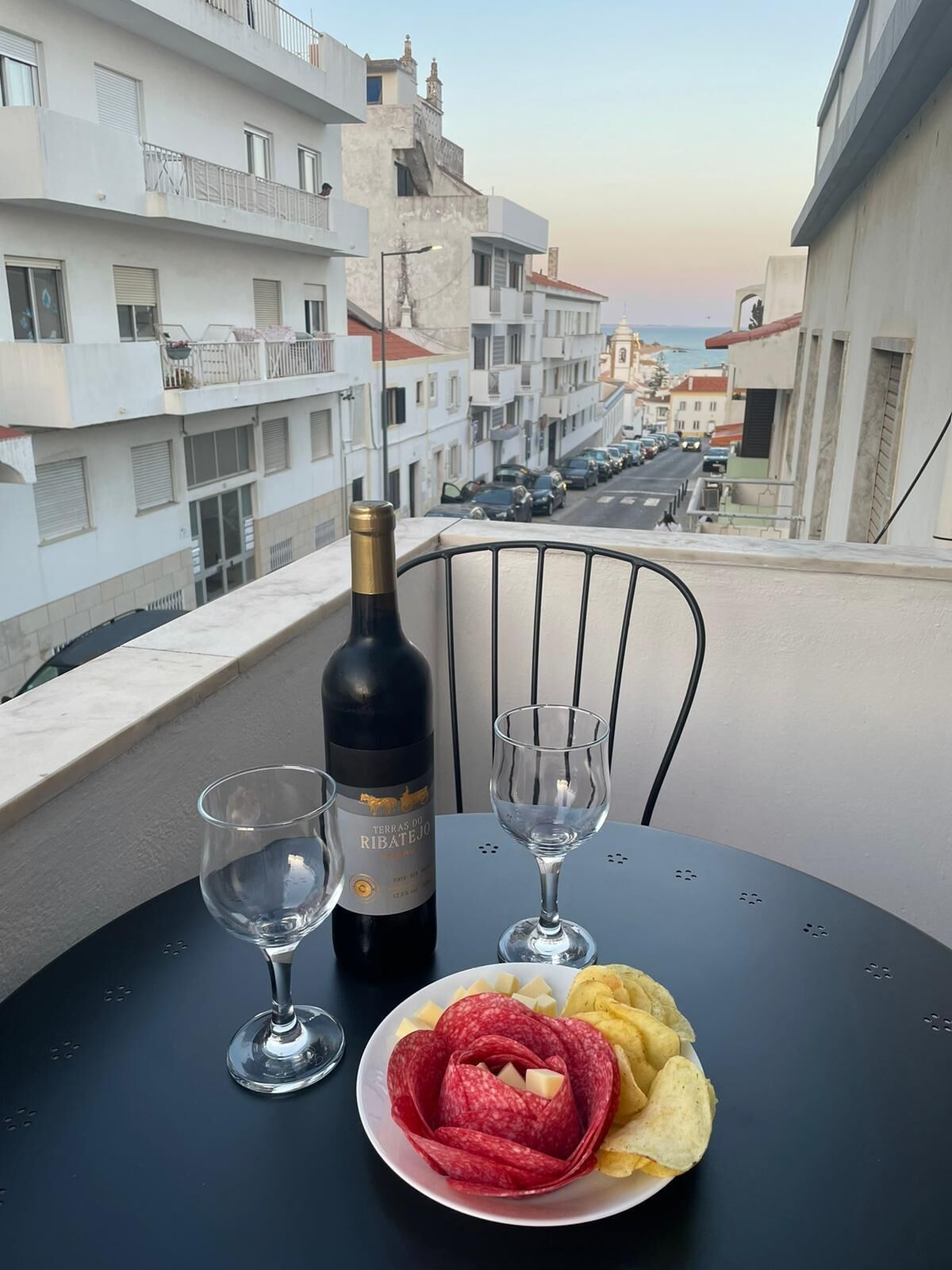 Ein Tisch mit einer Weinflasche, zwei Gläsern, einem Teller mit Salami und Chips sowie einem Stuhl.