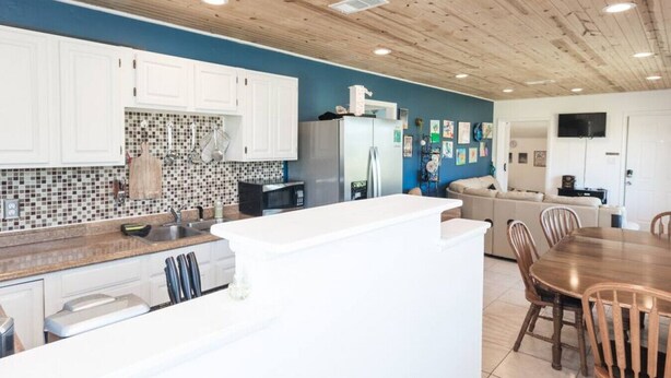 Een moderne keuken met witte kasten, een betegelde achterwand en een centraal eiland. Er is een eethoek met een houten tafel en stoelen.