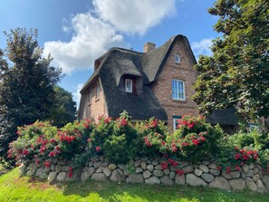 Ein traditionelles Haus mit einem steilen Satteldach, Ziegelwänden und einer Steinmauer mit blühenden Pflanzen davor.