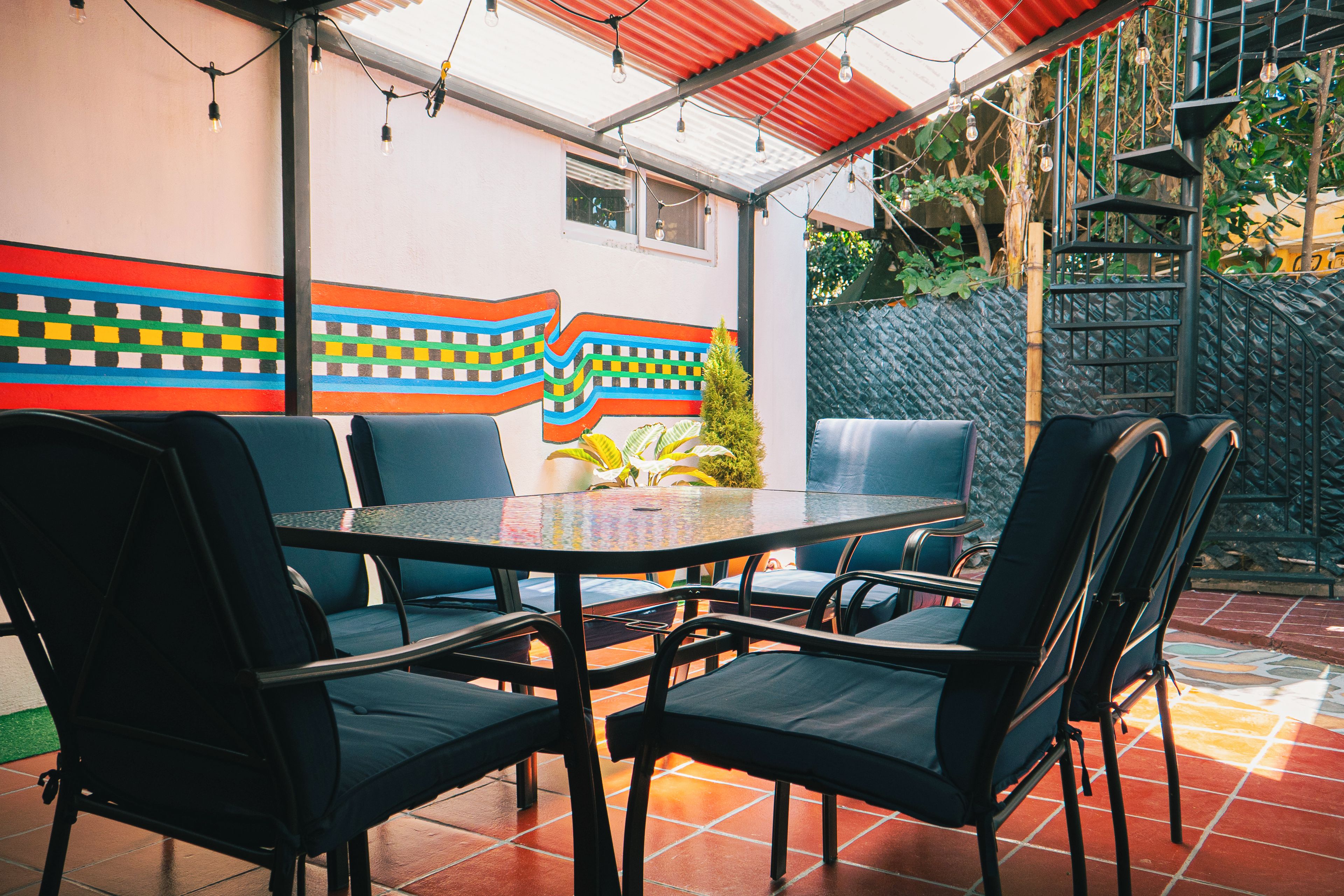 An outdoor seating area with a round table and chairs, a colorful mural on the wall, and a red roof structure.