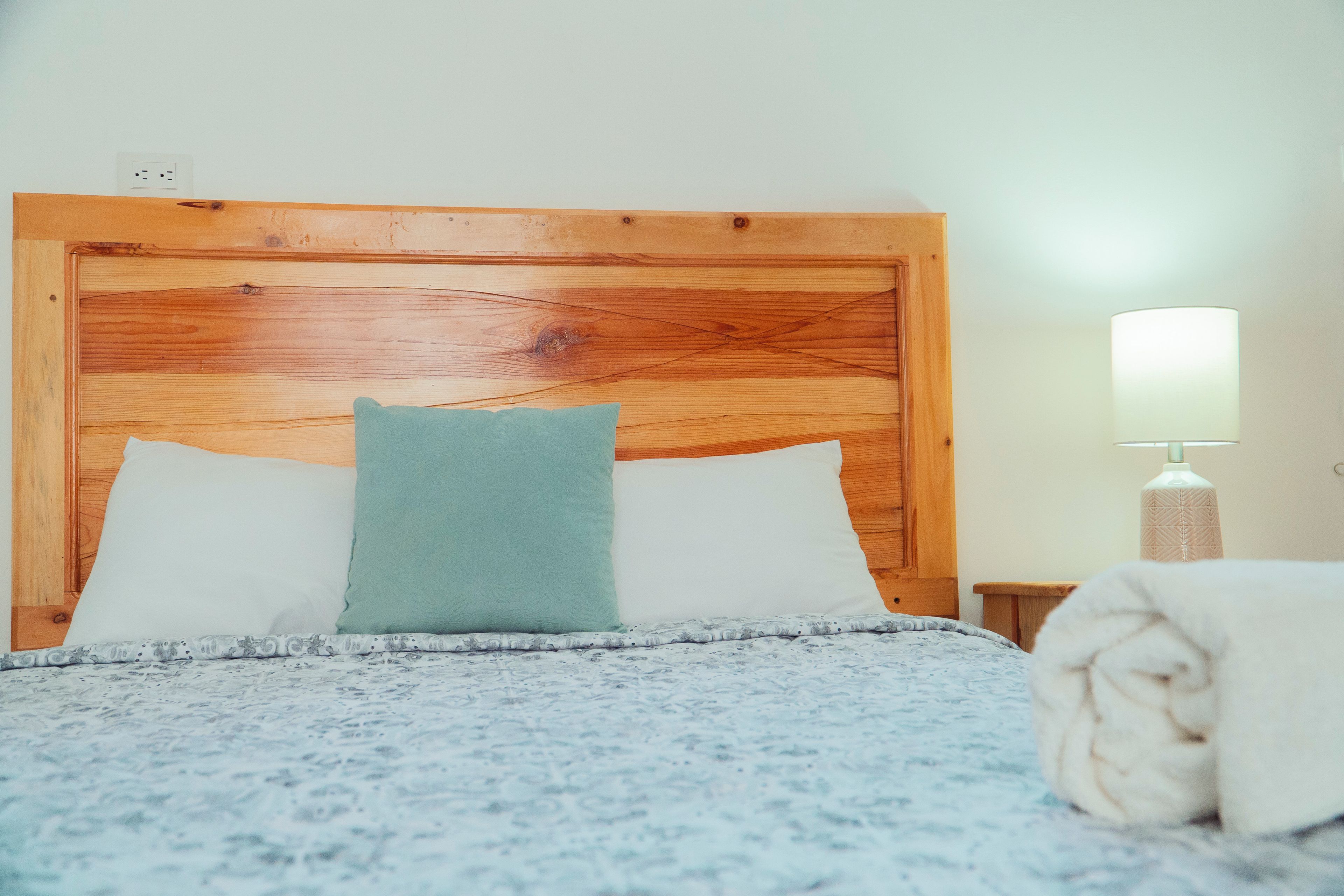 Un lit avec une tête de lit en bois, deux oreillers, une couverture, une lampe et une serviette enroulée.
