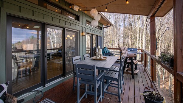 A covered outdoor deck with wooden furniture, string lights, and large windows offering views of a wooded area.