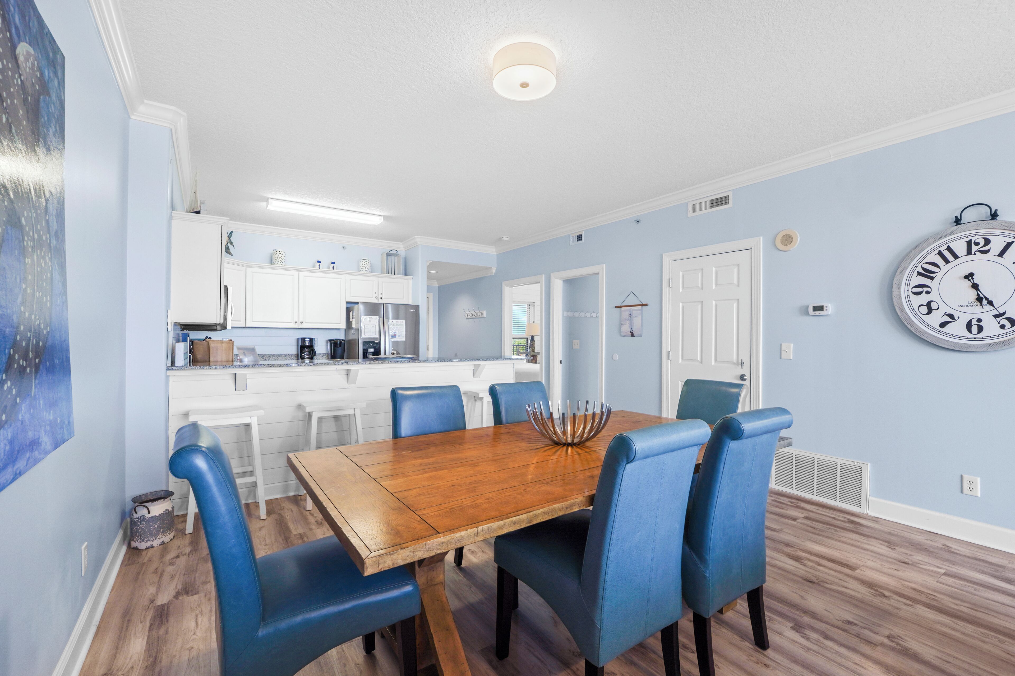 A dining area with a wooden table and blue chairs, a kitchen in the background, and a wall clock.