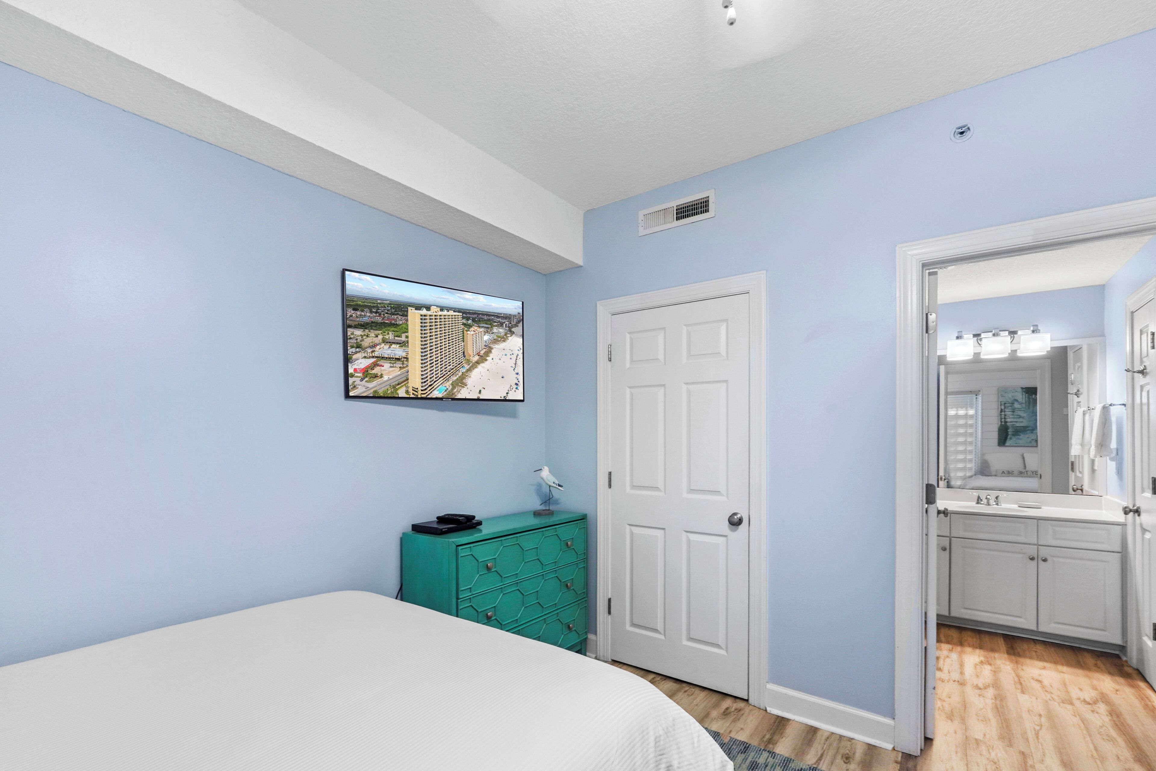 A bedroom with a bed, a teal dresser, a white door, and a bathroom visible through an open door.