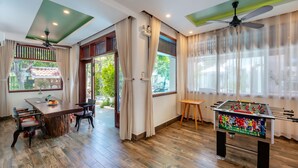 A room with a wooden floor, a foosball table, a dining table with chairs, and large windows with curtains.