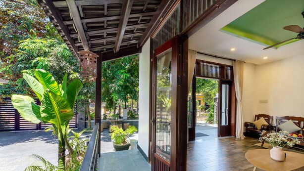 A spacious balcony with a view of lush greenery, a wooden door leading to a room with a sofa and a table, and a ceiling fan.