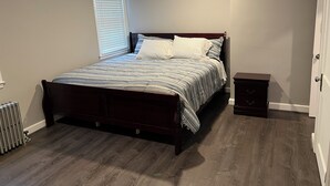 A bedroom with a wooden bed, a nightstand, a radiator, and a window with blinds. - Cozy 1-bedroom apartment with king size bed. (Towson)