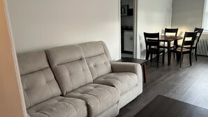 A living room with a beige sofa, a small table, and a dining area with chairs. - Cozy 1-bedroom apartment with king size bed. (Towson)