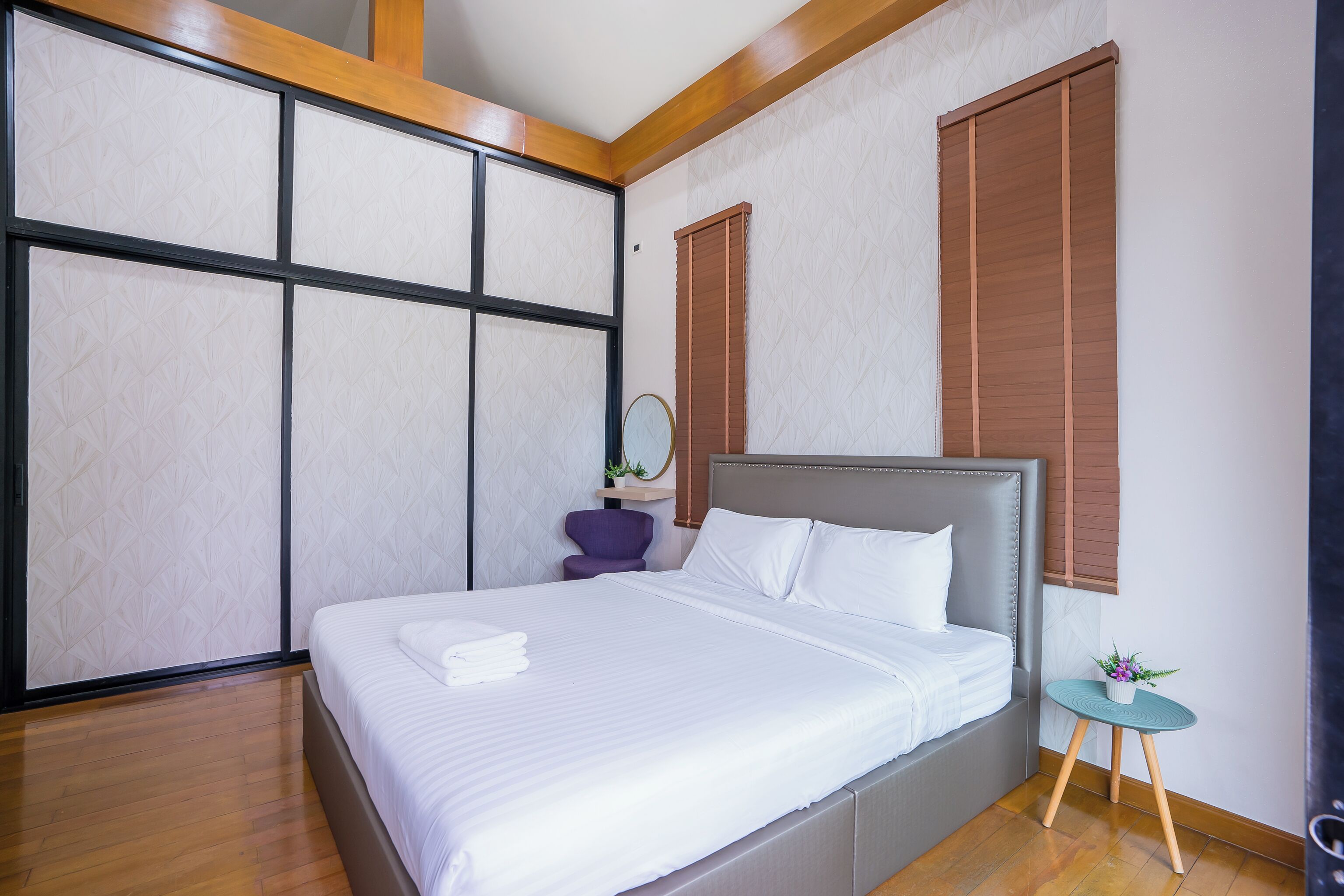 A neatly made bed with white linens, a small round table with a plant, and a purple chair in a modern room with wooden flooring and a large window with blinds.