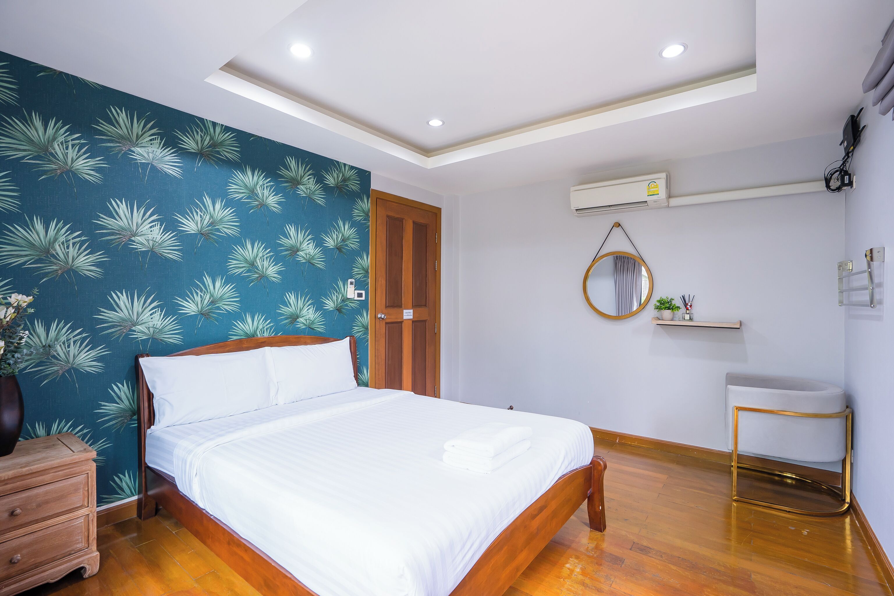 Una cama bien hecha con sábanas blancas, cabecera de madera y pared azul con estampado de hojas de palma. A la izquierda, un aparador de madera, un pequeño espejo redondo en la pared y un lavamanos blanco con marco dorado.