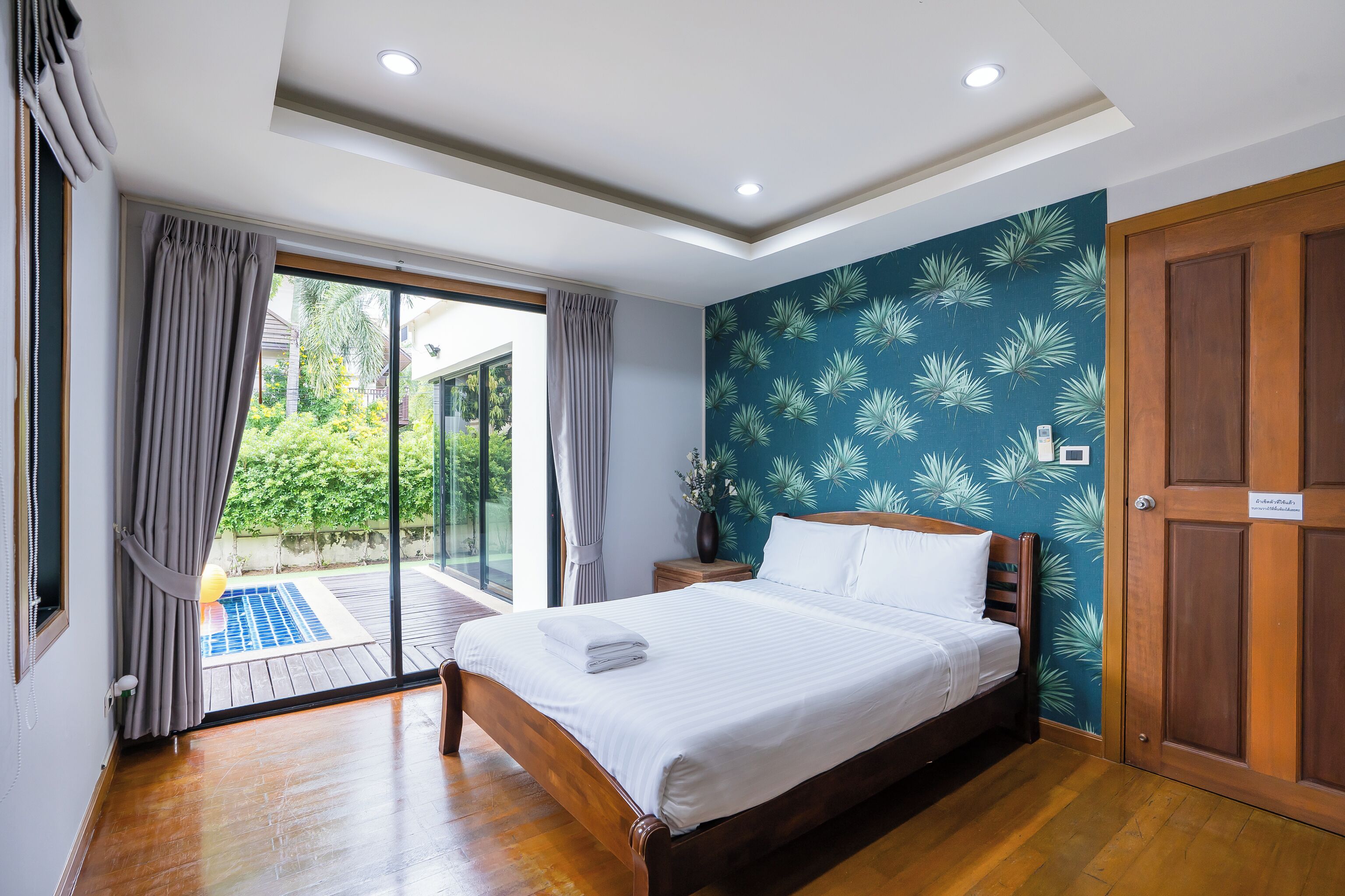 A bedroom with a wooden bed, a sliding glass door to a balcony, and a teal wall with a palm leaf pattern.
