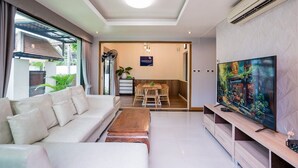 A modern living room with a sofa, coffee table, TV, and a dining area with chairs and a table. - Natcha Pool Villa by Warin (Pattaya)
