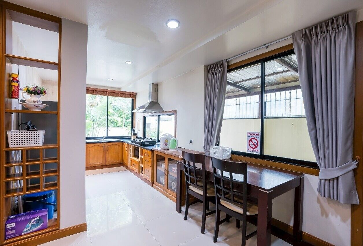 A kitchen with wooden cabinets, a dining table with chairs, a window with curtains, and a shelf with various items.