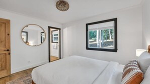 Een slaapkamer met een bed, een ronde spiegel, een raam met zonwering en een houten deur.