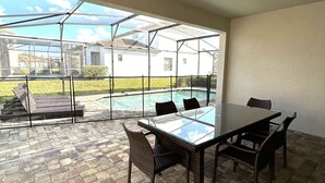 A covered patio with a glass table and chairs, a swimming pool, and a view of a house in the background. - Contemporary pool home, gated community, resort facilities! (Haines City)