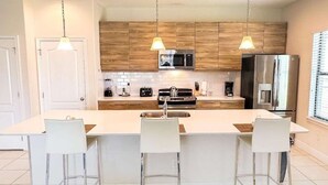 A modern kitchen with a central island, white bar stools, wooden cabinets, a stainless steel refrigerator, and pendant lighting. - Contemporary pool home, gated community, resort facilities! (Haines City)
