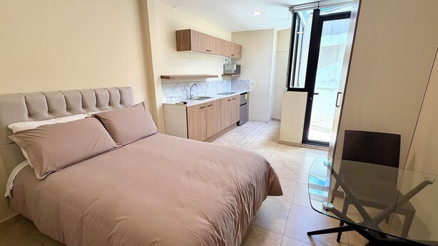 Une chambre avec un lit, une table en verre et une vue sur une cuisine et un balcon.