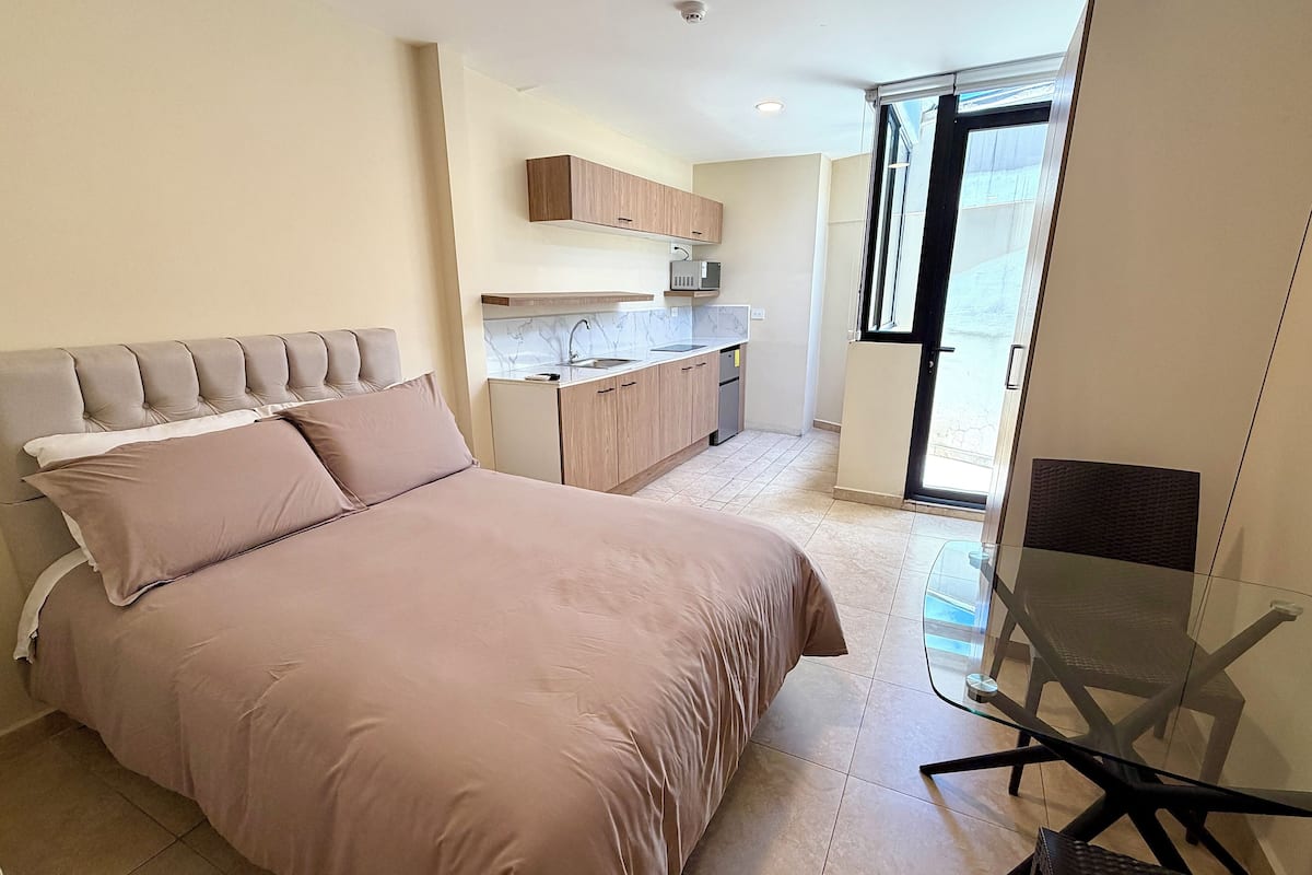 A bedroom with a bed, a glass table, and a view of a kitchen and balcony.