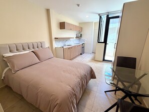A bedroom with a bed, a glass table, and a view of a kitchen and balcony.