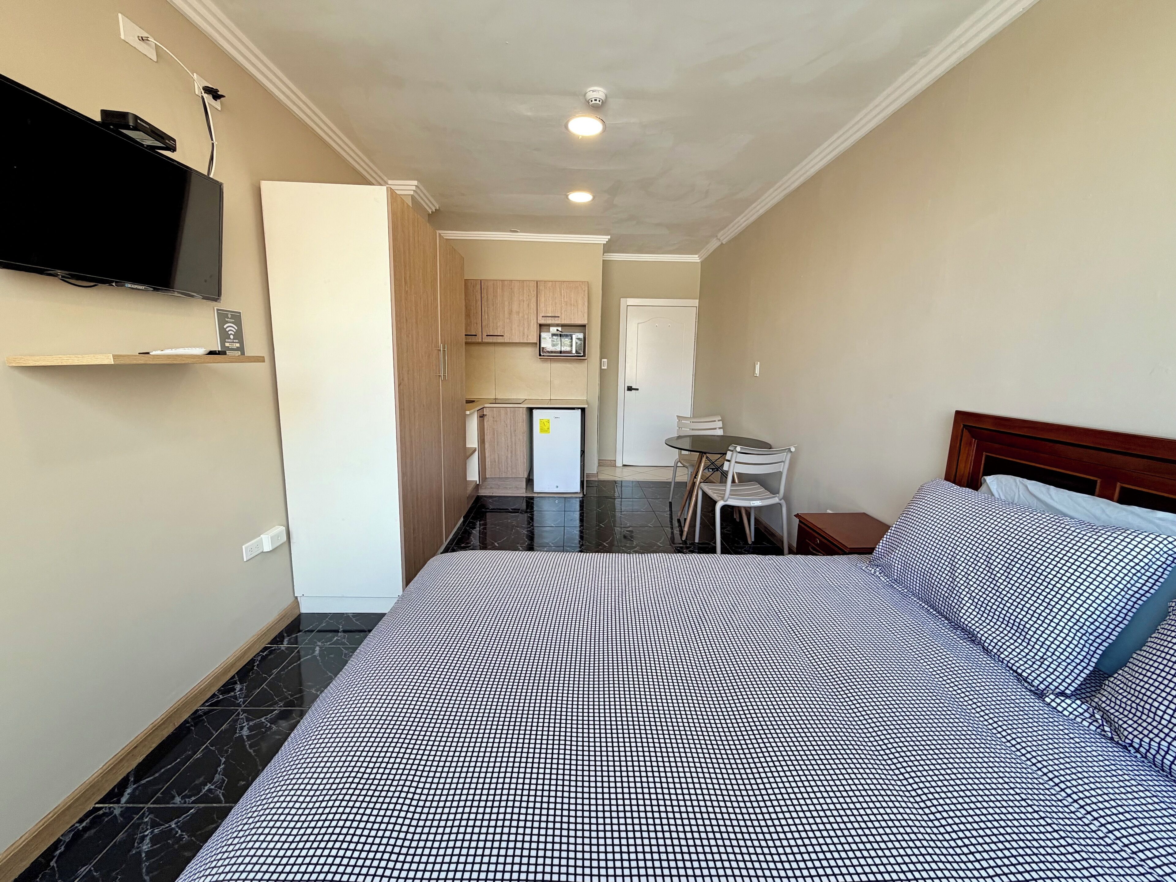 A hotel room with a bed, a TV mounted on the wall, a small table with chairs, and a kitchenette in the background.