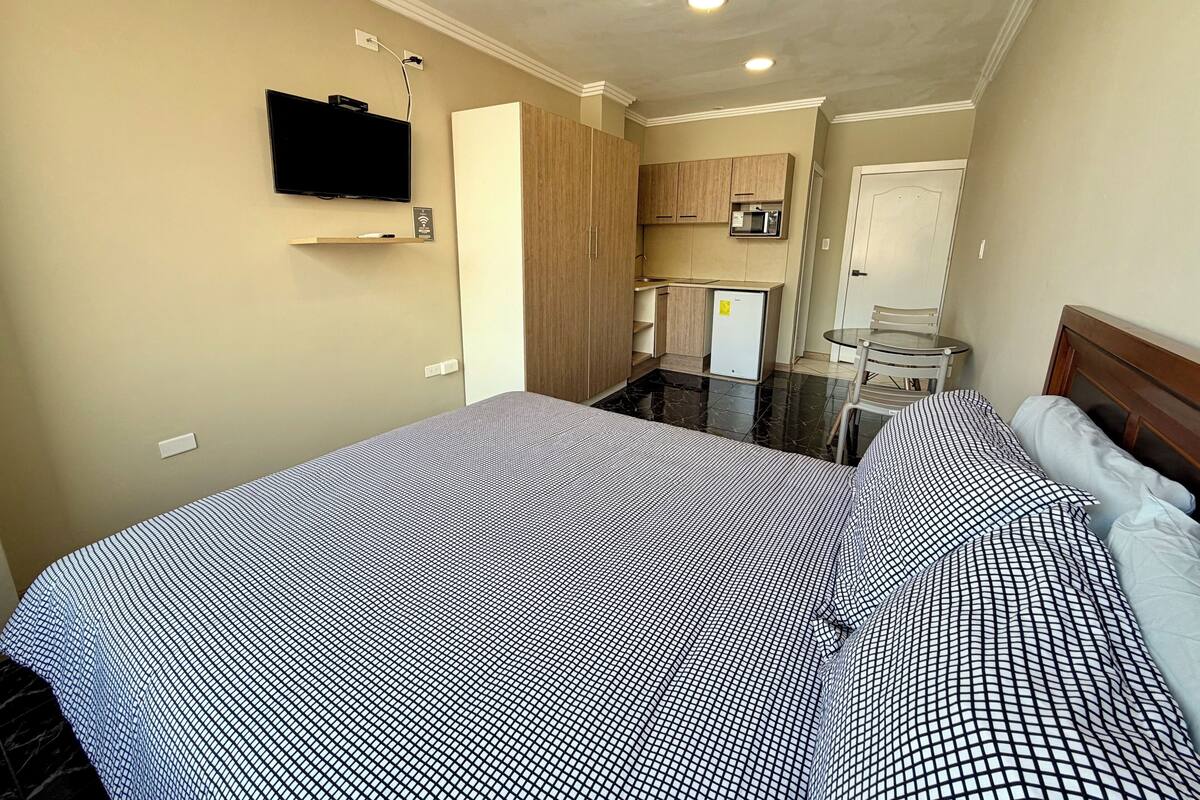 A hotel room with a bed, a TV mounted on the wall, a small kitchenette, and a glass table with a chair.