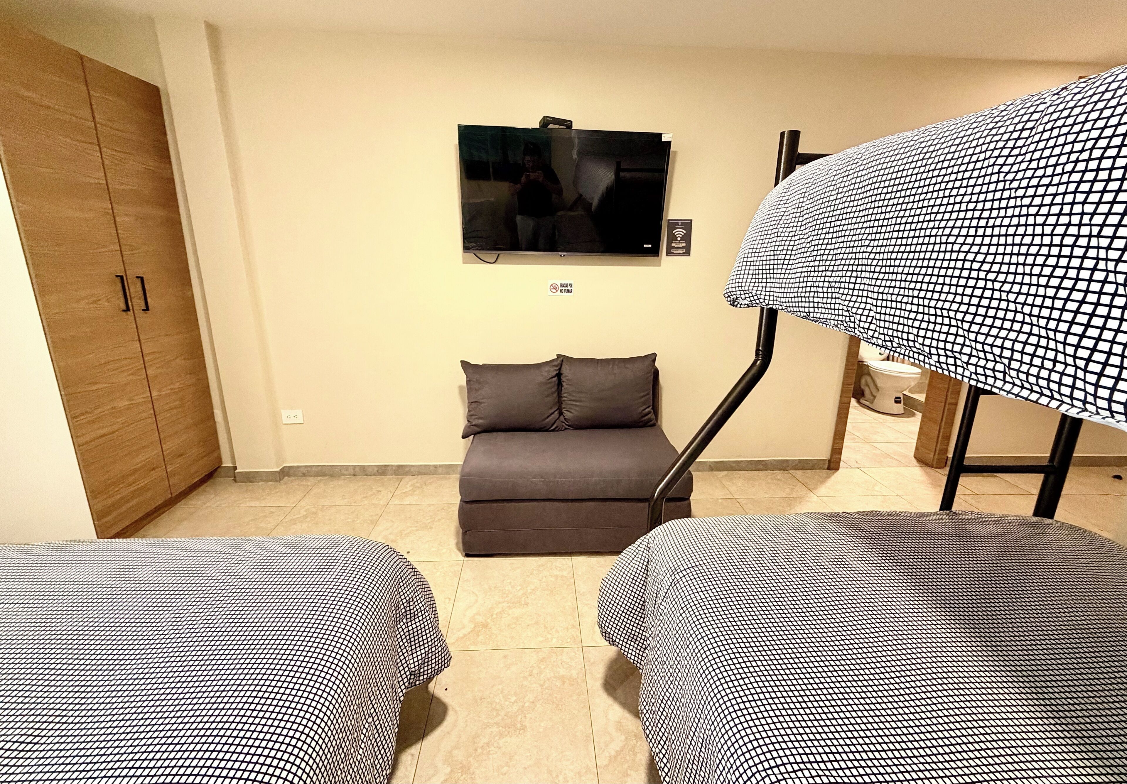 Quarto com sofá, duas camas com colchas xadrez, televisão na parede e guarda-roupa de madeira.