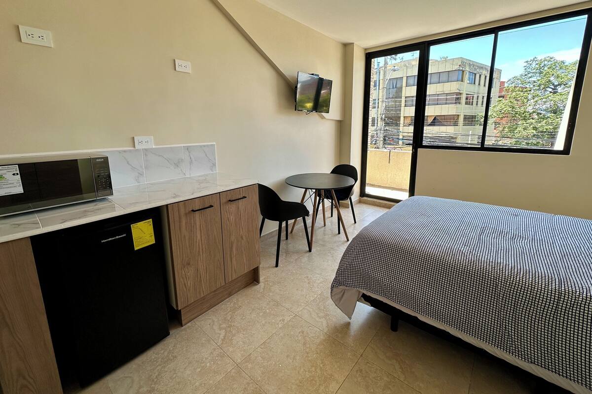 A compact hotel room with a bed, a small kitchenette, a dining area, and a large window offering a city view.