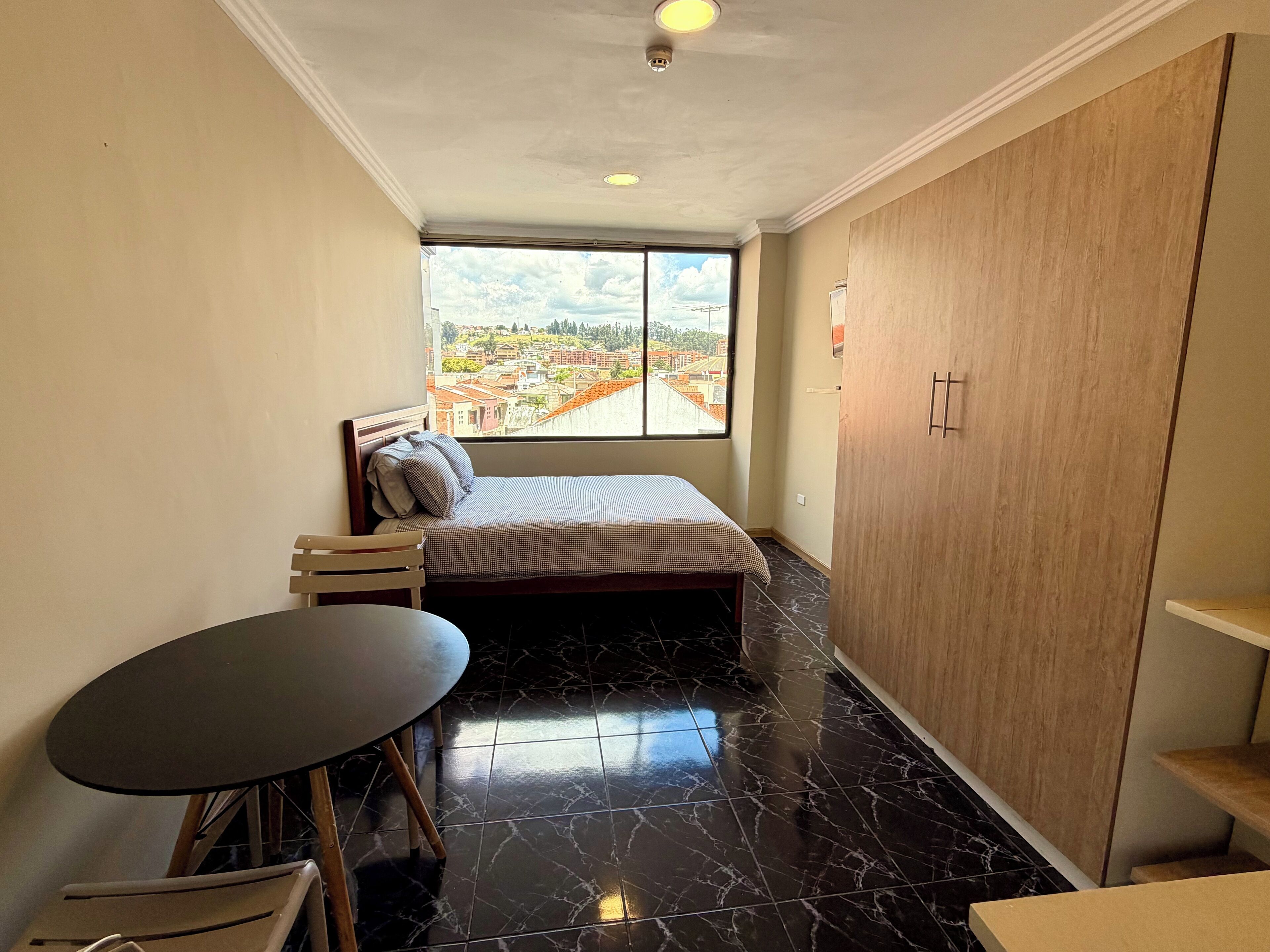 Una habitación pequeña y funcional con una cama, una mesa y vista a un paisaje urbano a través de un ventanal amplio.