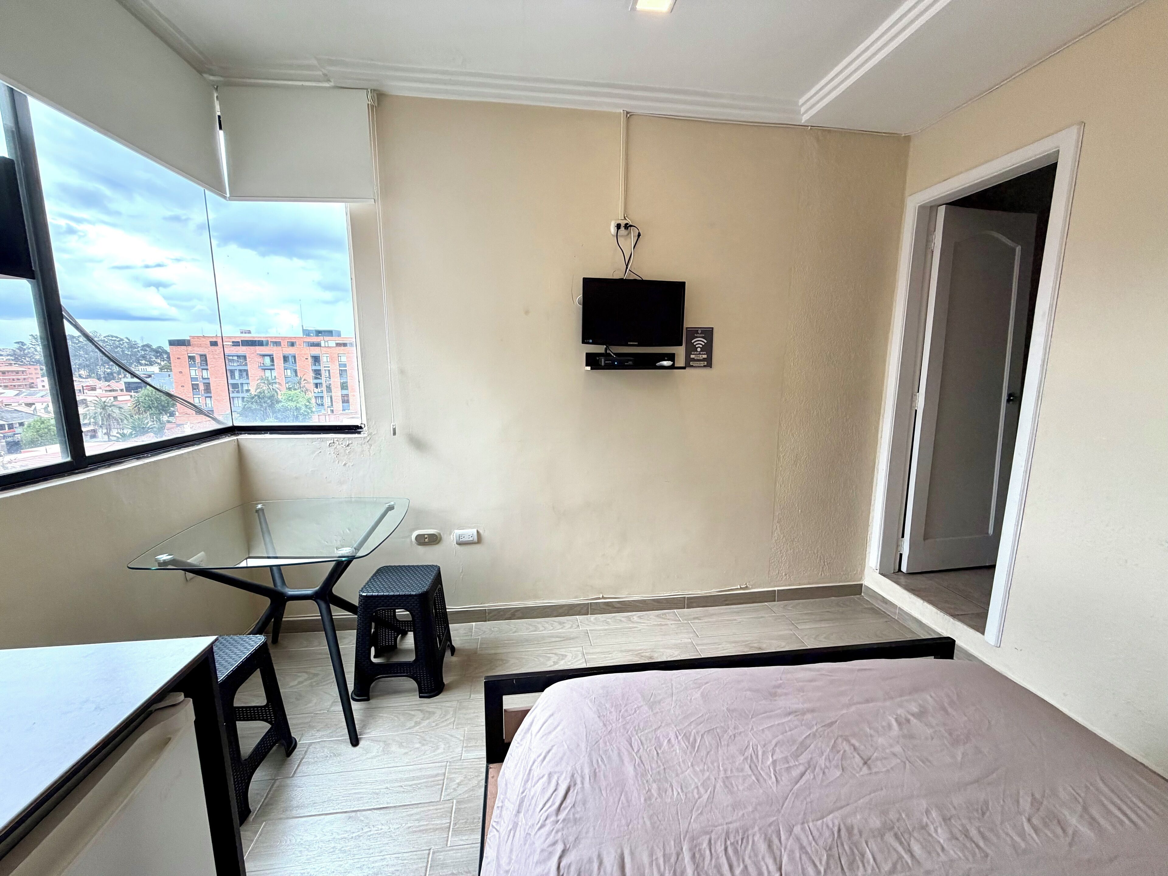 A small, modern room with a glass desk, a black stool, a bed with a white cover, a TV mounted on the wall, and a view of buildings through a window.