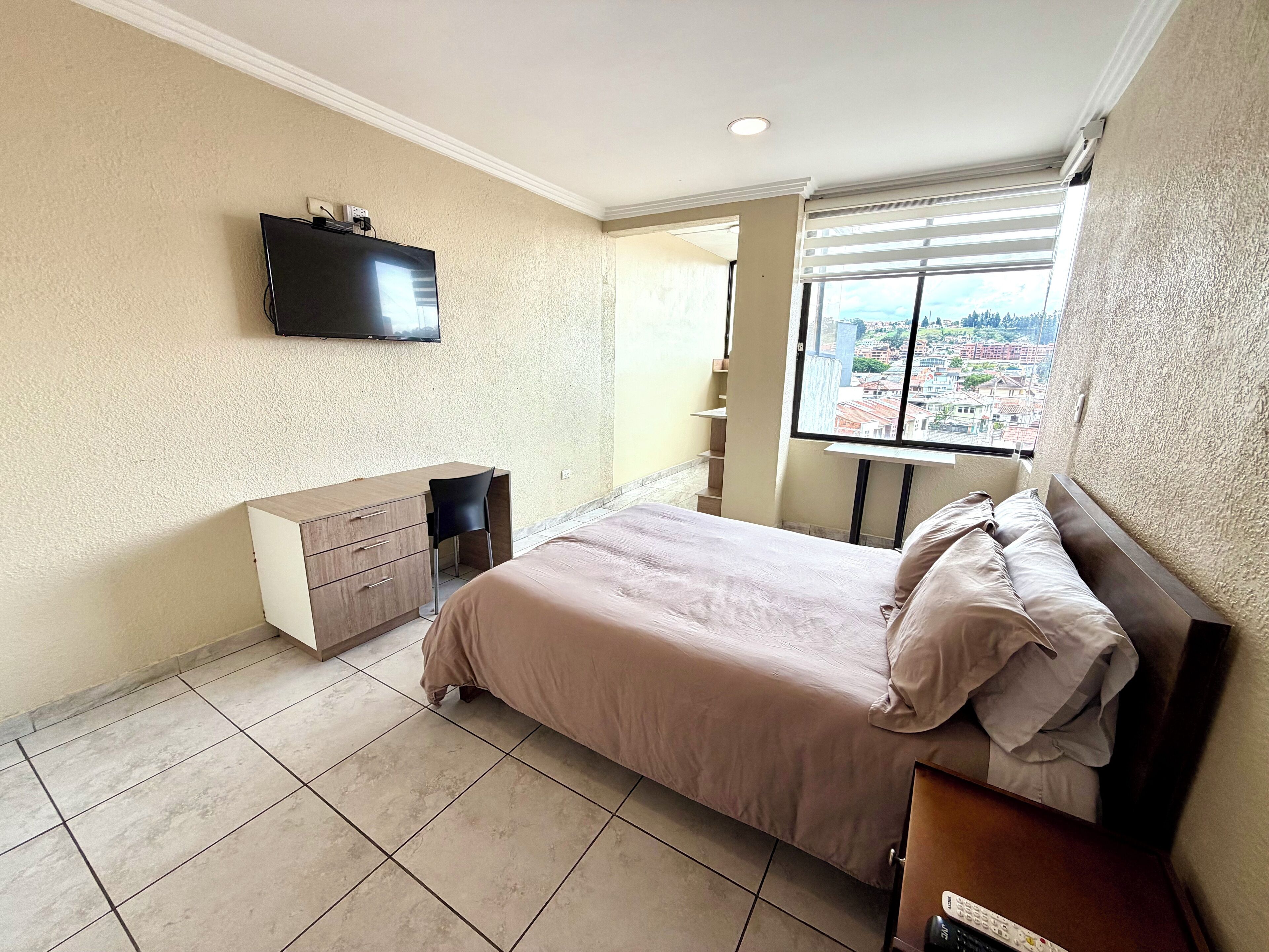 A bedroom with a bed, a desk, a chair, a TV mounted on the wall, and a window with a city view.