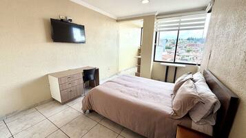 A bedroom with a bed, a desk, a chair, a TV mounted on the wall, and a window with a city view.