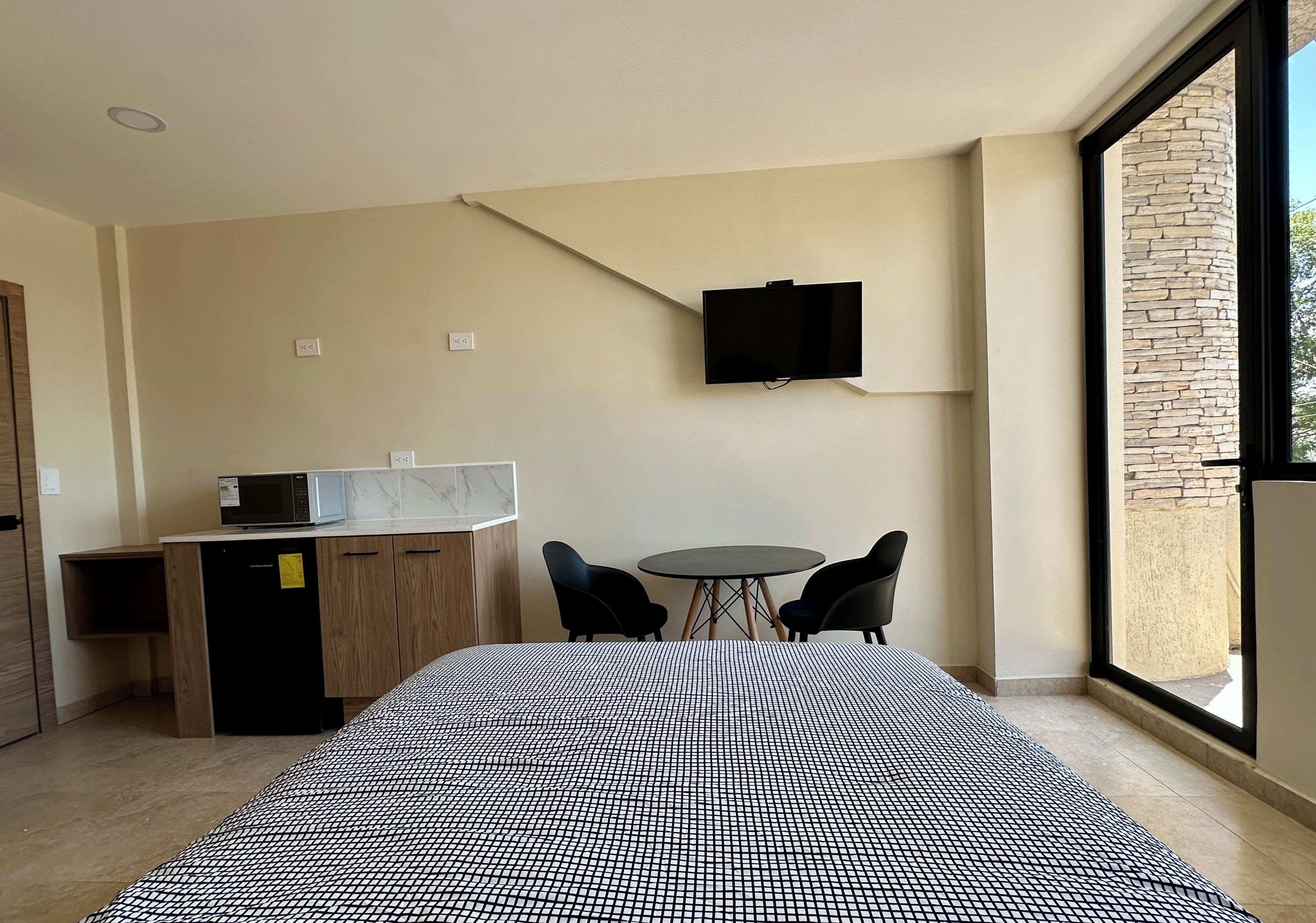 A hotel room with a bed, a desk with a microwave, a TV on the wall, two chairs, and a large window.