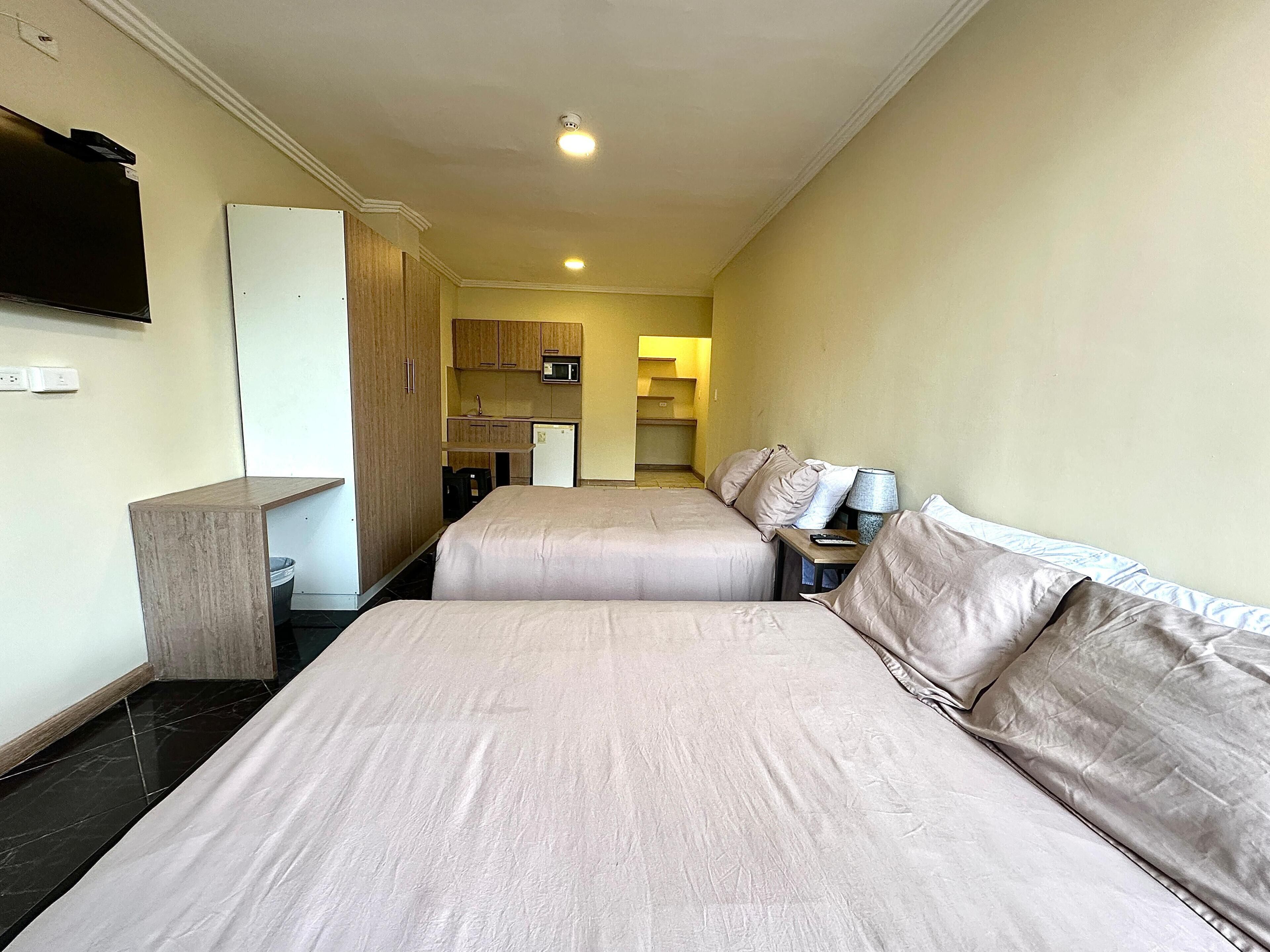 Quarto de hotel com duas camas, cozinha compacta, micro-ondas e televisão na parede.