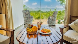 A wooden table with a basket of fruits and a plate of sliced fruit, two floral-patterned chairs, and a view of greenery and mountains through a window. - Guldaar - A Luxury Forest Retreat (Rajgarh)