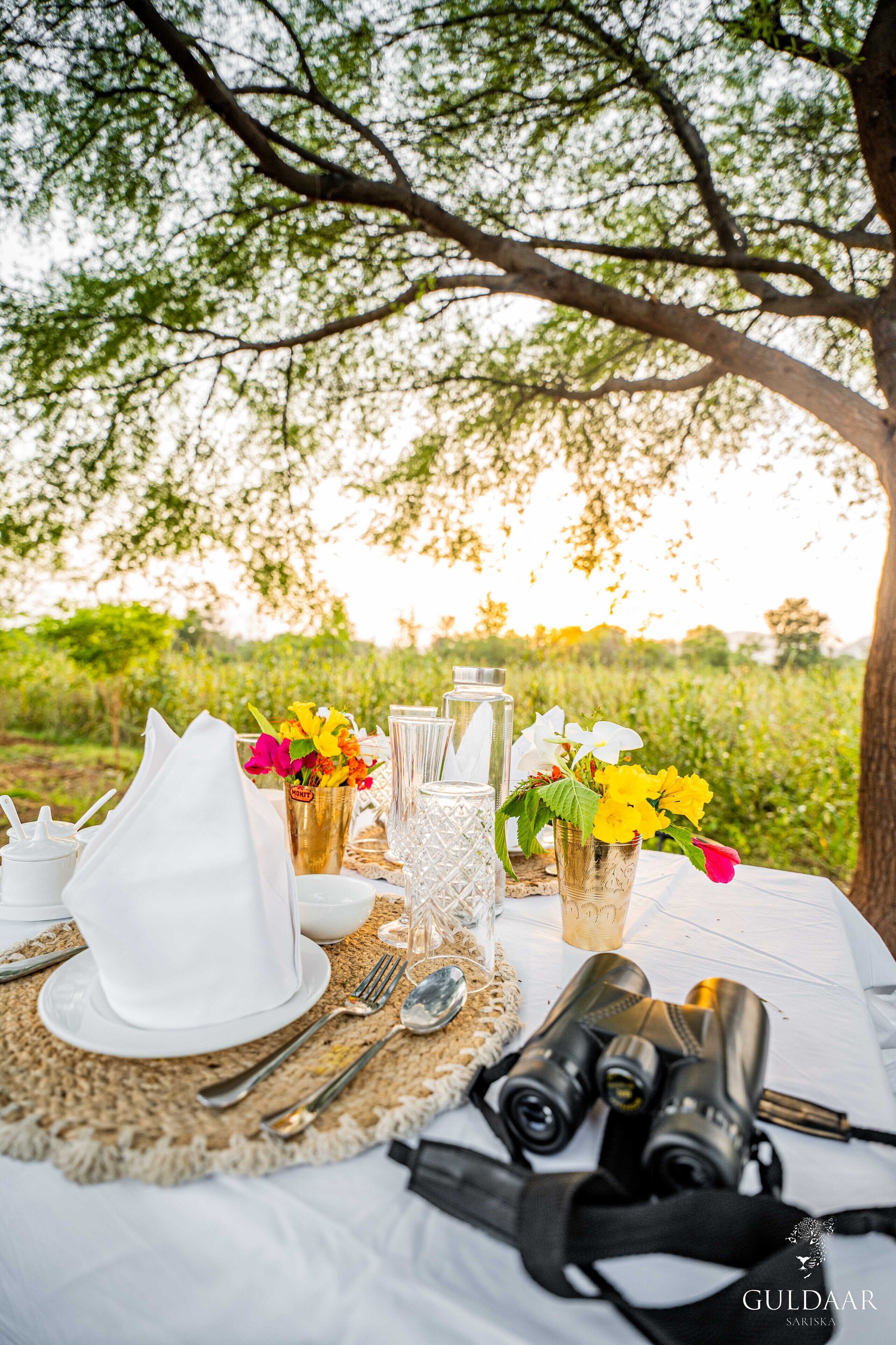 Una mesa colocada al aire libre con vistas a árboles y follaje, que incluye un mantel blanco, cubiertos, vasos, una cámara y un florero con flores.