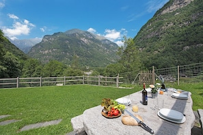 A table set outdoors with a mountain view, a bottle, a plate with food, and a knife. - Rustico alla bolla (Sonogno)