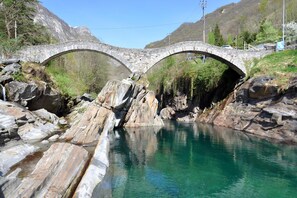 A stone bridge over a clear river with rocky banks and mountains in the background. - Rustico alla bolla (Sonogno)