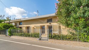 Ein zweistöckiges Gebäude mit beiger Fassade, einem Metallzaun und einem kleinen Garten mit rosa Blumen.
