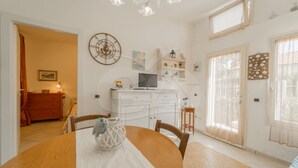 Ein Essbereich mit einem runden Tisch, Stühlen, einer weißen Tischdecke, einer Dekoschüssel, einer Uhr, einem Fernseher und einem Fenster mit Vorhängen.