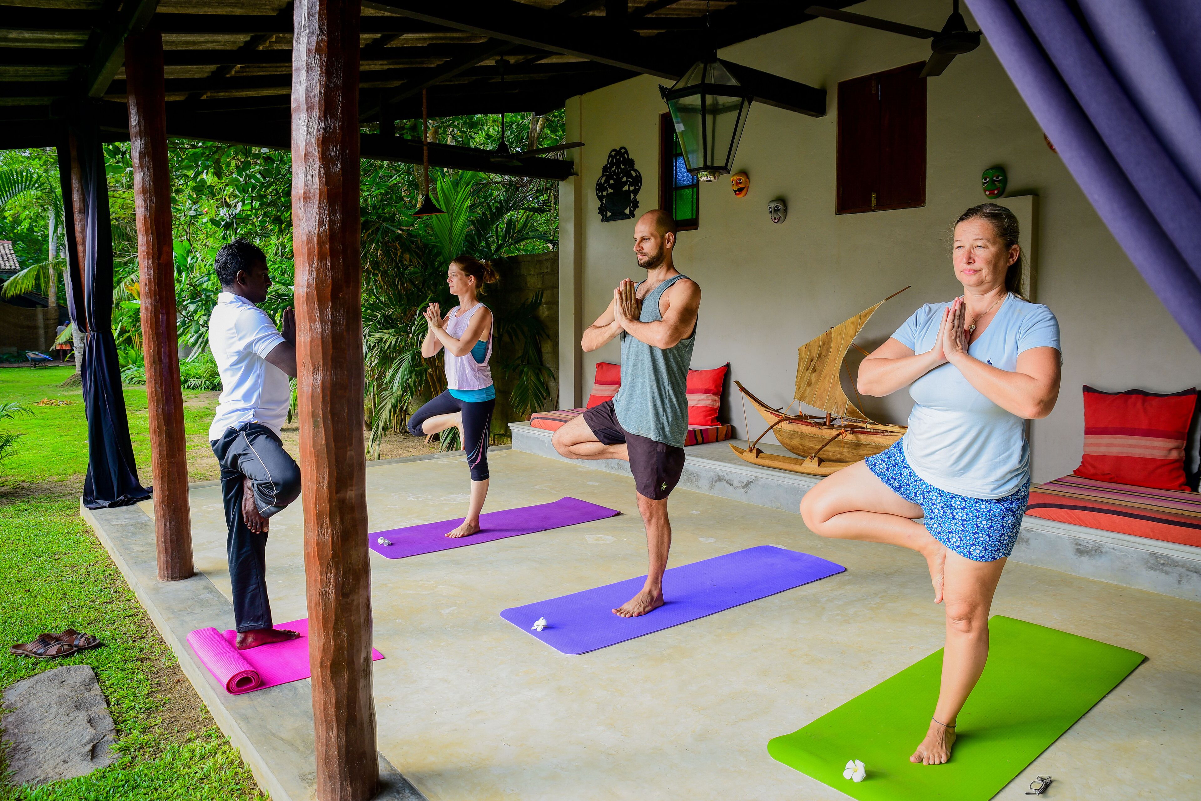 Aula de yoga em área externa coberta, com pilares de madeira, tapetes verdes e um sofá com almofadas vermelhas.
