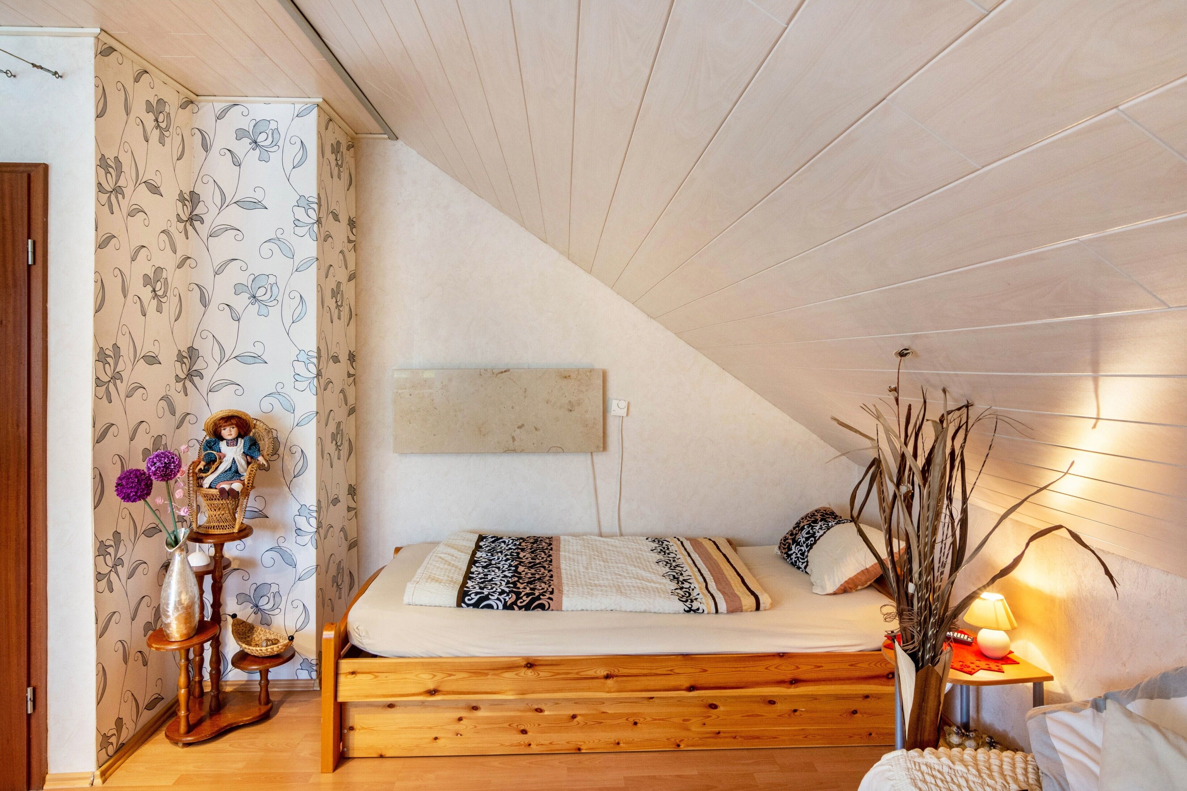 A bedroom with a sloped ceiling, a bed with patterned bedding, a wooden dresser, and a vase with flowers.