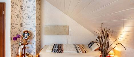 A bedroom with a sloped ceiling, a bed with patterned bedding, a wooden dresser, and a vase with flowers.