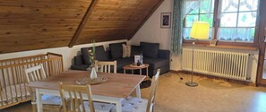 Et hyggeligt rum med trælofter, et spisebord med stole, en sofa med puder, et lille sofabord, en gulvlampe og et vindue med blonde gardiner.