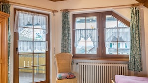 Una habitación con cama, una silla de mimbre con cojín, una ventana con cortinas de encaje y un radiador.
