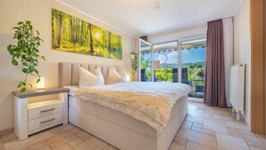 A bedroom with a bed, a nightstand, a potted plant, a painting, and a view of greenery through a sliding door.