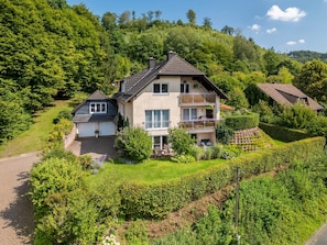 A two-story house with a balcony, surrounded by lush greenery and a hilly landscape.