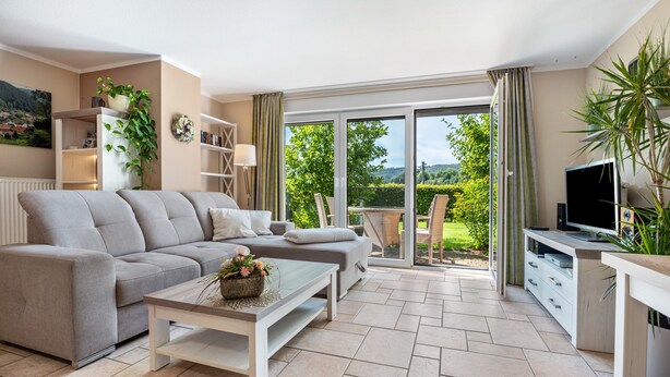 A living room with a sofa, coffee table, and TV stand, a balcony with a view of greenery, and a bookshelf.