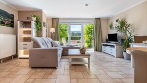 A modern living room with a sofa, coffee table, TV, and a view of greenery through large windows.