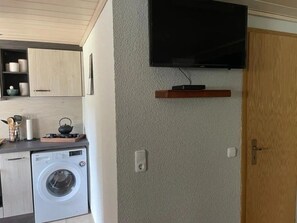 Une cuisine compacte équipée d’un lave-linge, d’un téléviseur à écran plat fixé au mur et d’une porte en bois.