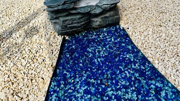 A rock waterfall feature with a blue glass base, surrounded by gravel and a brick wall. - Apartment 'Bella Petit' with Private Terrace and Wi-Fi (Büdingen (Wolferborn))